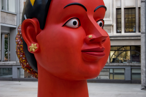 A monumental large scale public sculpture depicting the head of a deity painted red and adorned with jewellery and a hair piece.