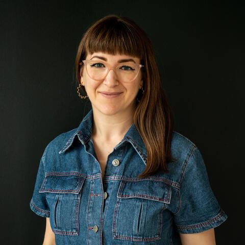 Portrait of artist Ellie Shipman, who is a white presenting female with long brown hair, wearing glasses and a dark blue denim shirt. She is smiling and the photo is set against a black background. 