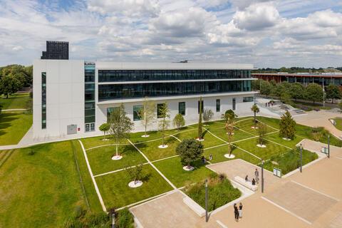 An image of landscaped trees set on a university campus that features a large rectangular white building and a courtyard.